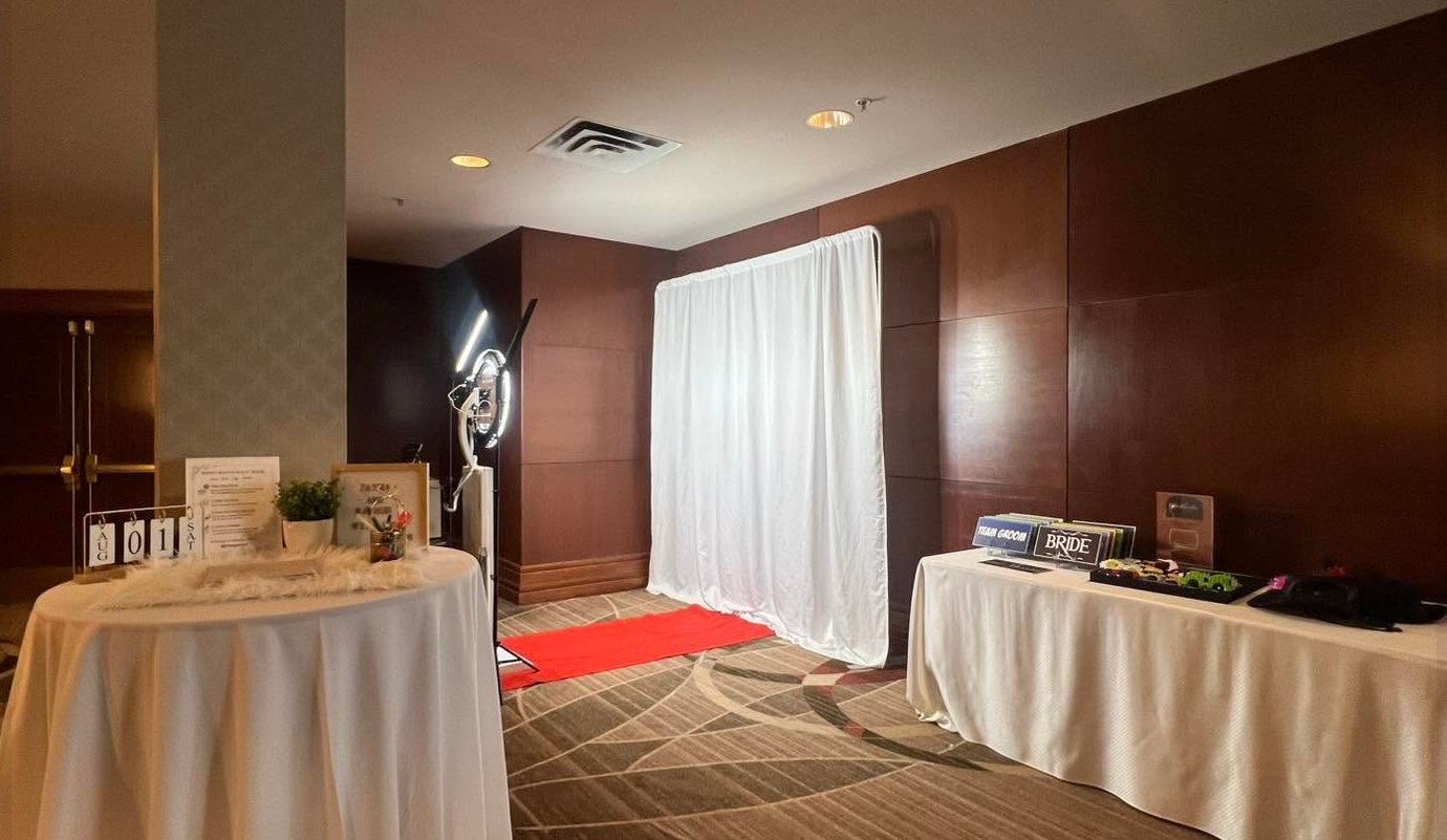 Photograb ring-light booth set up at a hotel wedding, with a white backdrop, red carpet, a welcome table and a table of Team Bride and Team Groom props